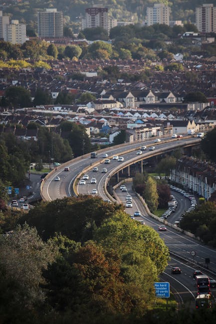 Aerial view of a multi-lane road on a curved section, with vehicles including cars and vans in motion during daylight hours. The road passes through a suburban area with tightly packed terraced houses
